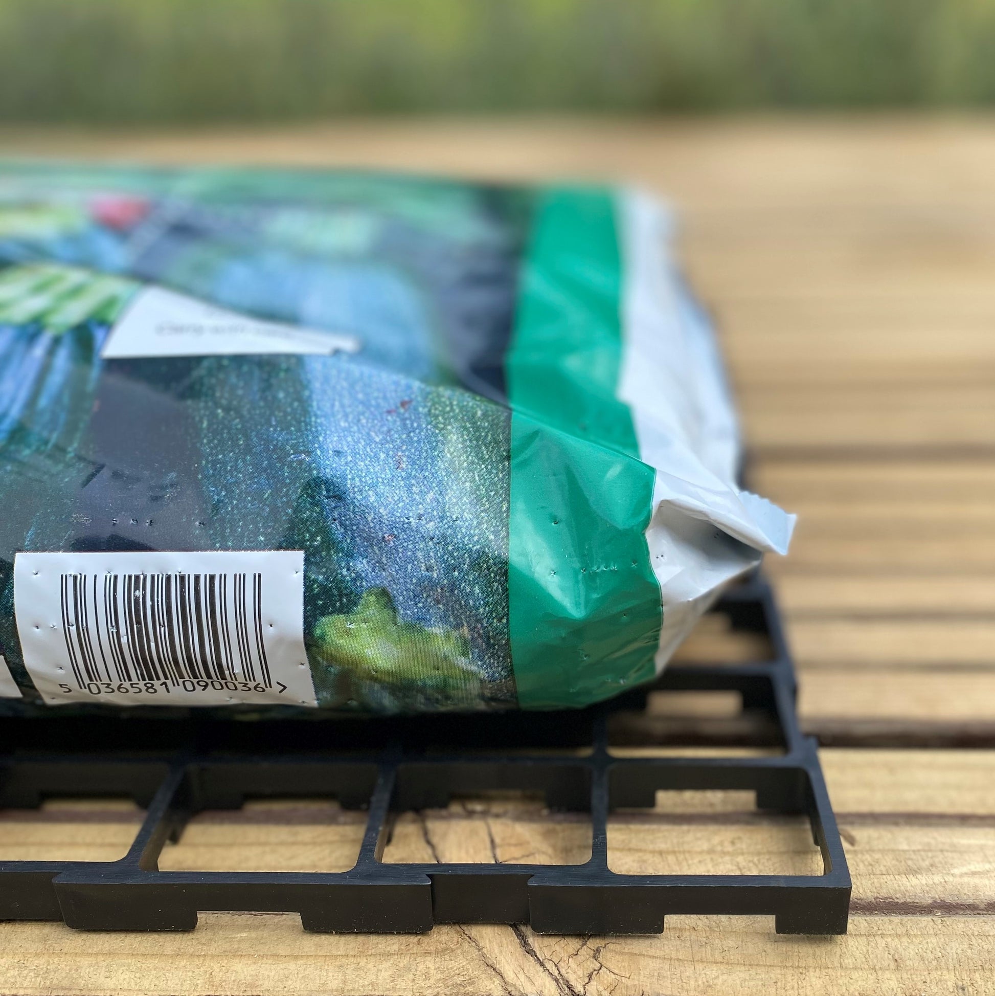 A black interlocking growbag mat placed under a green growbag, set on a wooden surface.