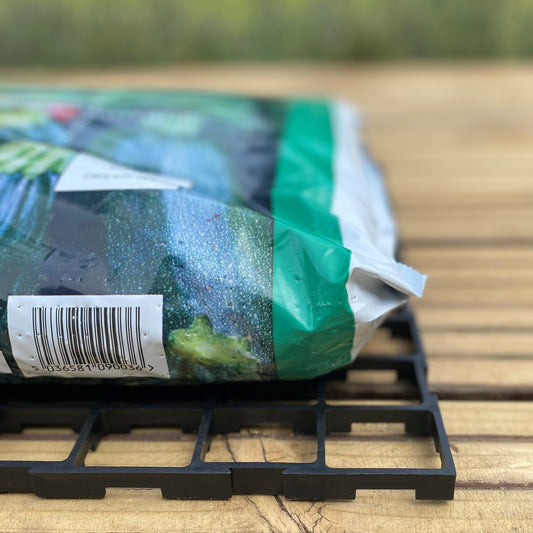 A black interlocking growbag mat placed under a green growbag, set on a wooden surface.