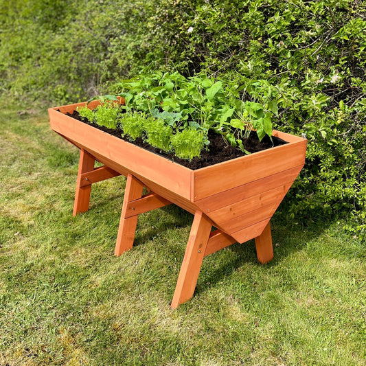 A large wooden raised vegetable bed planter placed on grass, filled with soil and green plants.