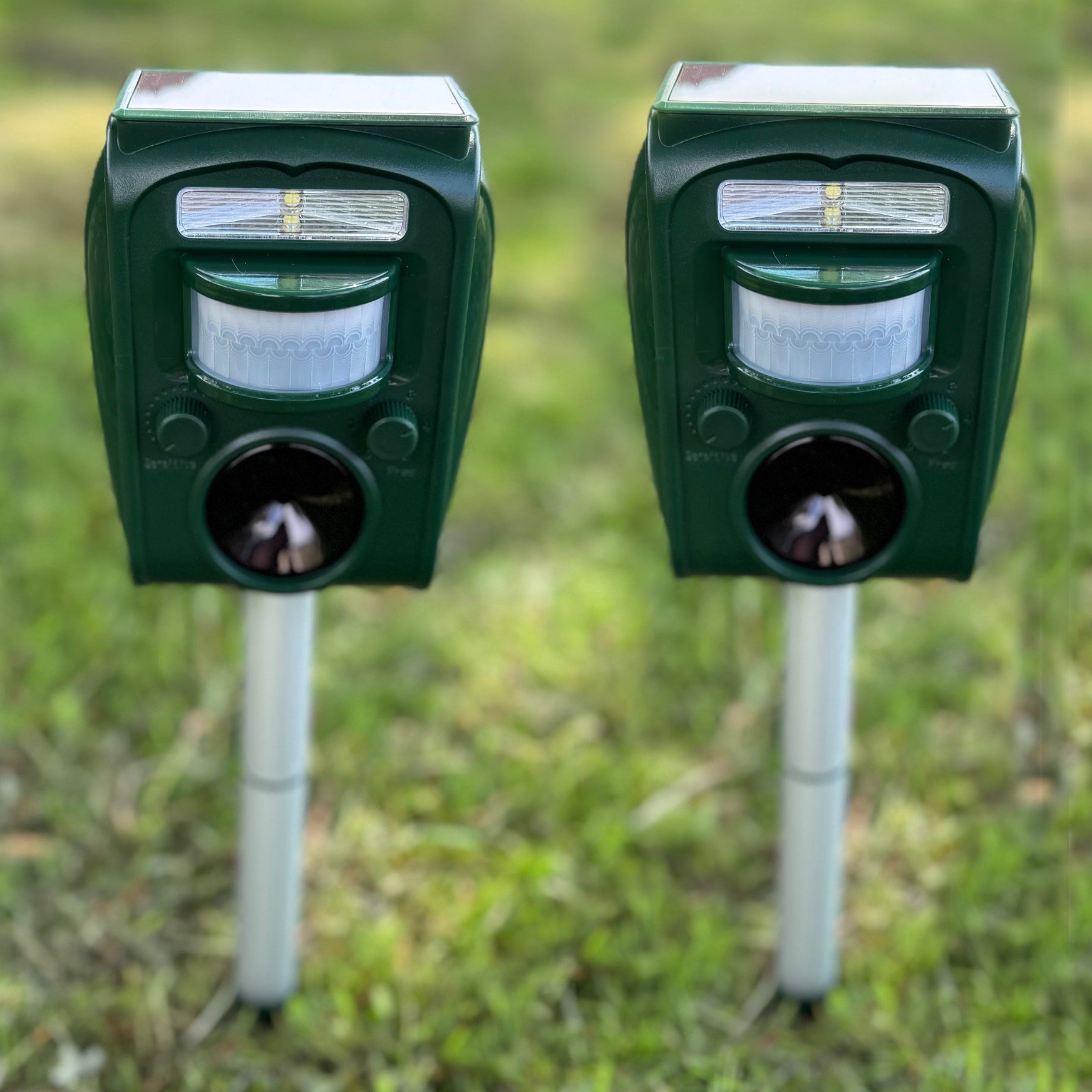 A set of two green solar powered ultrasonic repellers mounted on silver stakes in a grassy area.