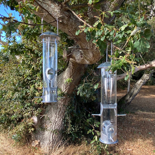 Two clear aluminium bird feeders with multiple feeding ports hanging from a tree.