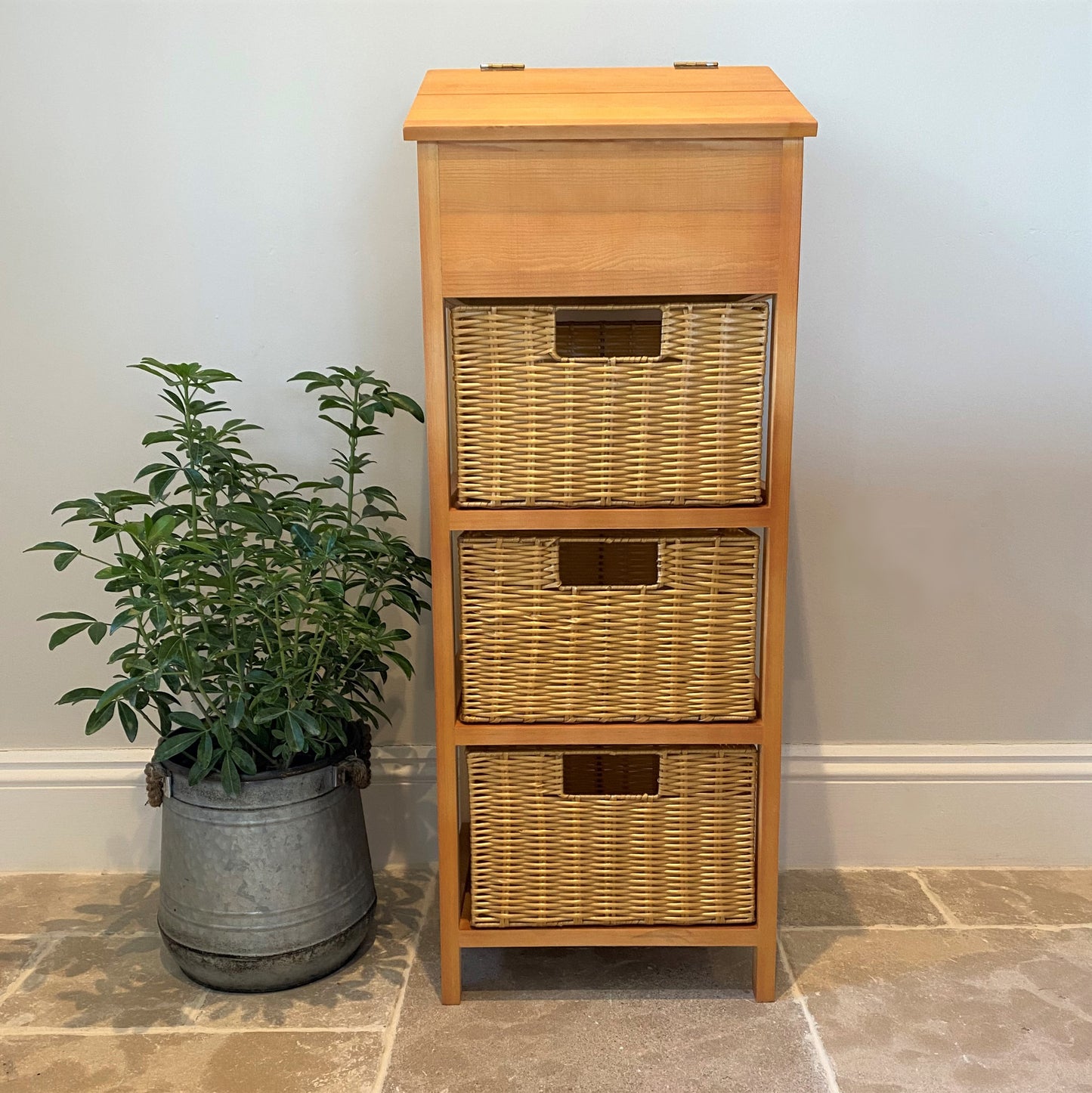 A wooden vegetable and potato storage unit with three wicker style baskets, placed next to a potted plant.