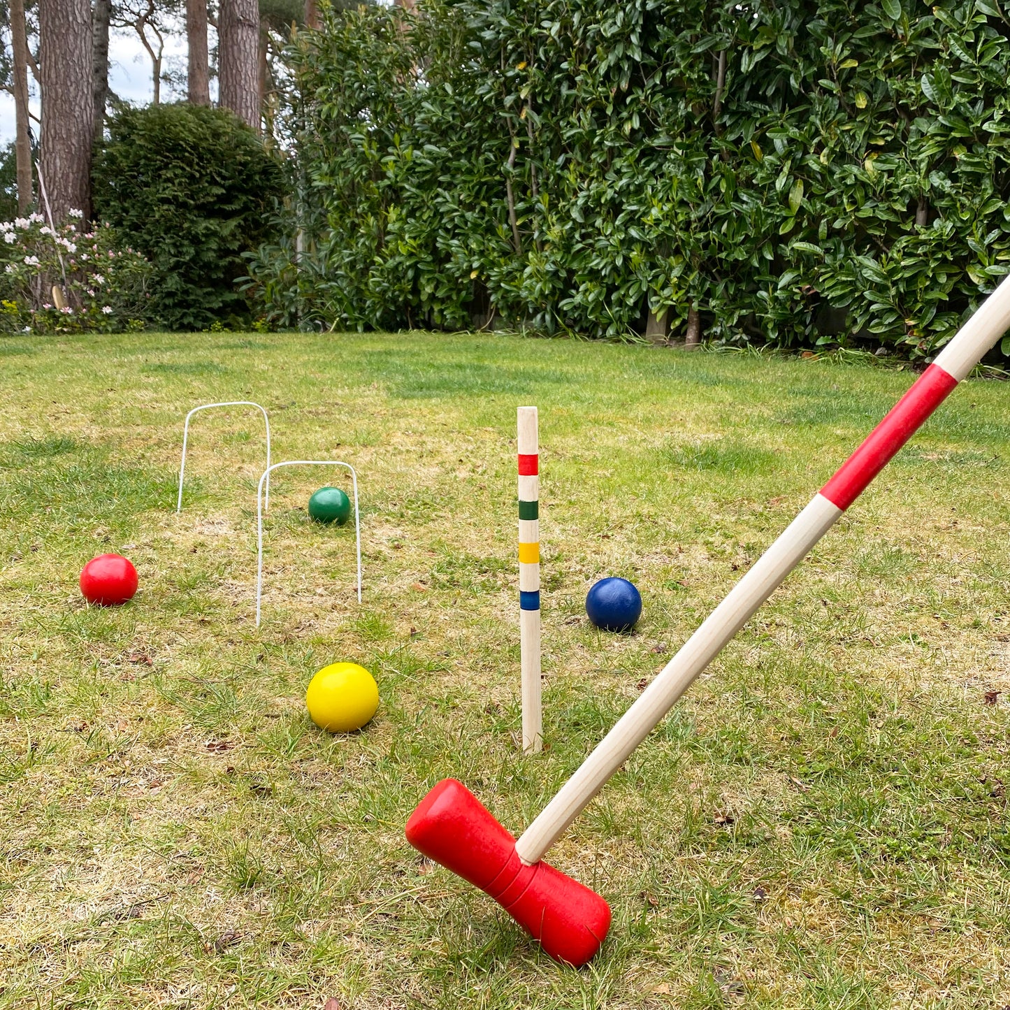 A wooden croquet set with red, blue, and yellow balls, along with a mallet, set up on a lawn.