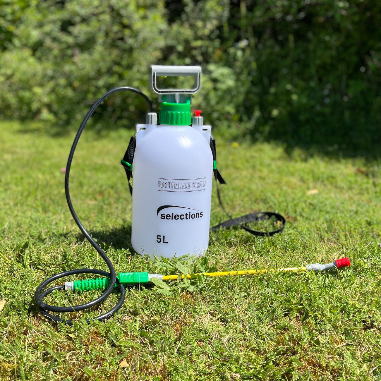 A white 5-litre garden shoulder pressure sprayer with green and red accents, including a shoulder strap, hose, and spray lance, placed on grass.