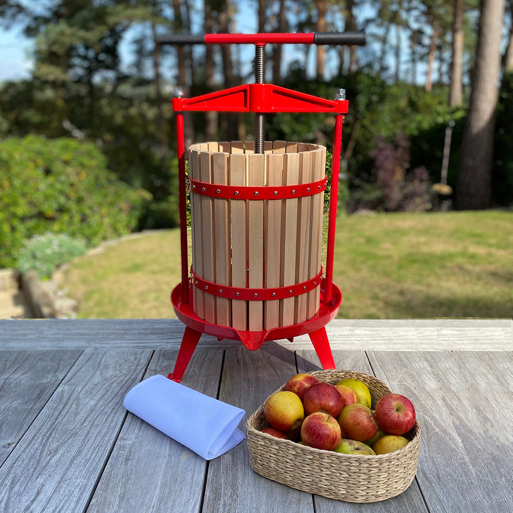 A traditional 18 litre fruit press with a red frame and a T handle, alongside a basket of apples and a cloth on a wooden surface.