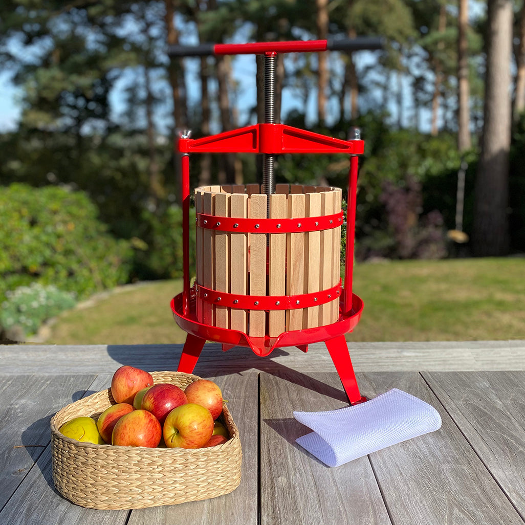 A traditional 12 litre fruit press with a red frame and a T handle, alongside a basket of apples and a piece of paper on a wooden surface.