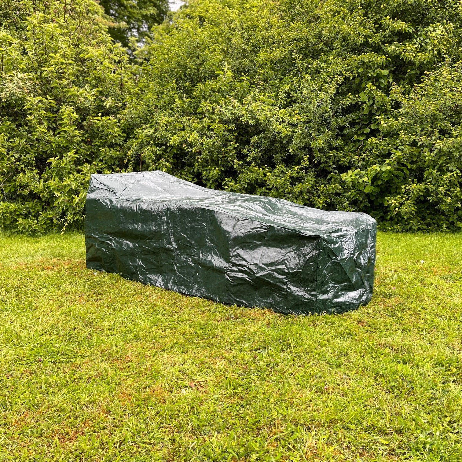 A green waterproof sunbed cover placed on a garden lounger, with trees and shrubs in the background.