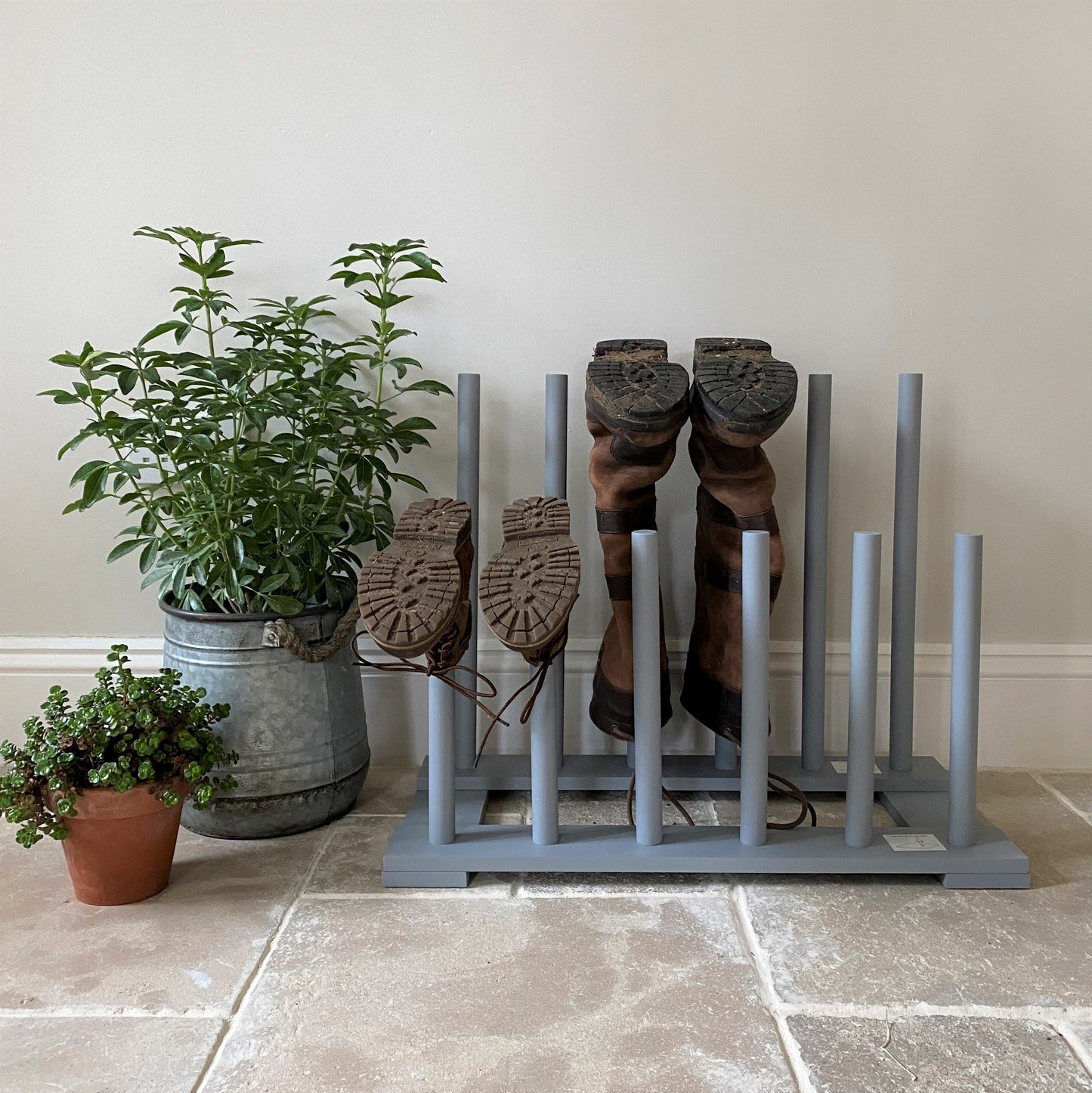 A wooden wellington boot rack in light grey, accommodating three pairs of boots, placed on a tiled floor next to a potted plant and a small bucket.