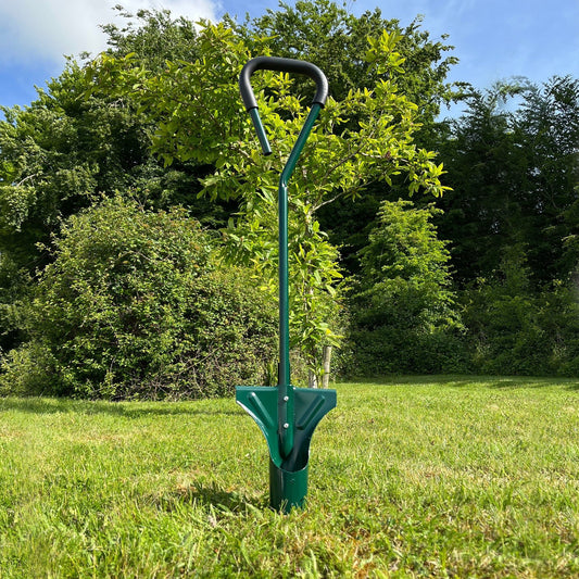 A green long handled metal garden bulb planter standing upright on a grassy field with trees in the background.