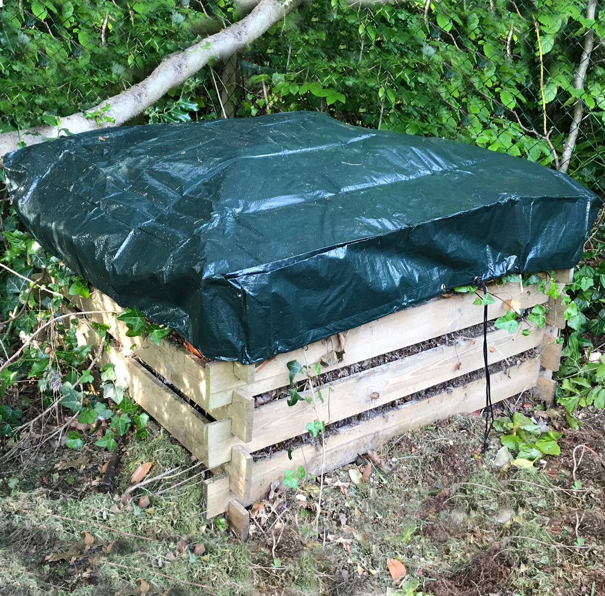 A green polyethylene cover placed on a wooden slatted composter in an outdoor setting.