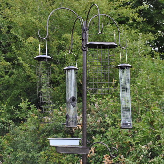 A metal bird feeding station with four large feeders hanging from a pole, set against a backdrop of trees. The station also includes a water dish and a mealworm tray.