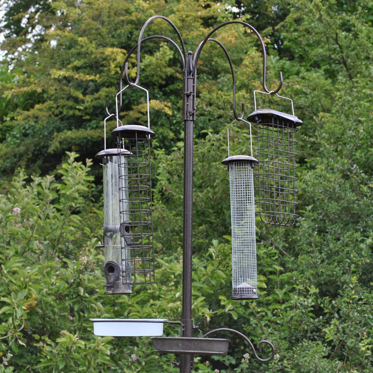 A metal bird feeding station with four large feeders hanging from a central pole, set against a backdrop of trees.