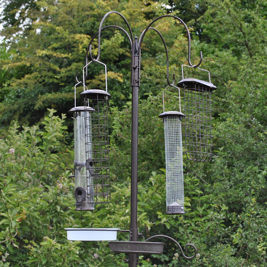 A metal bird feeding station with four large feeders hanging from a central pole, set against a backdrop of trees.