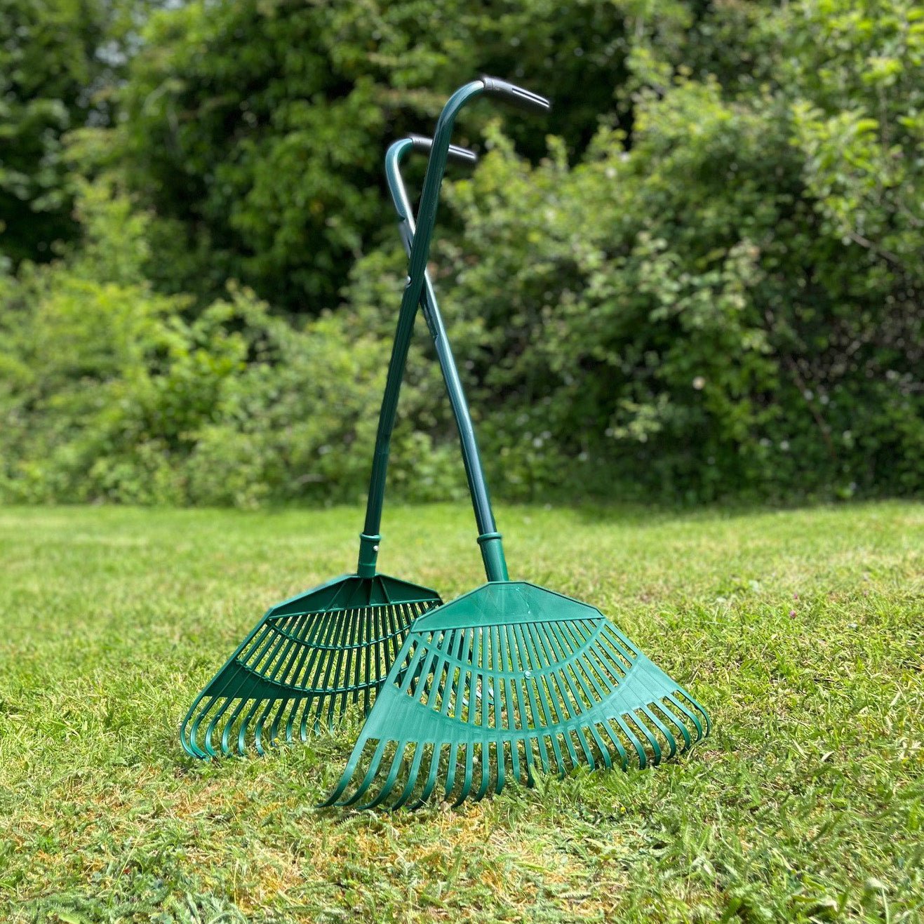 Two green long handled leaf collecting grabs with tubular steel frames and plastic grip handles, standing on grass.