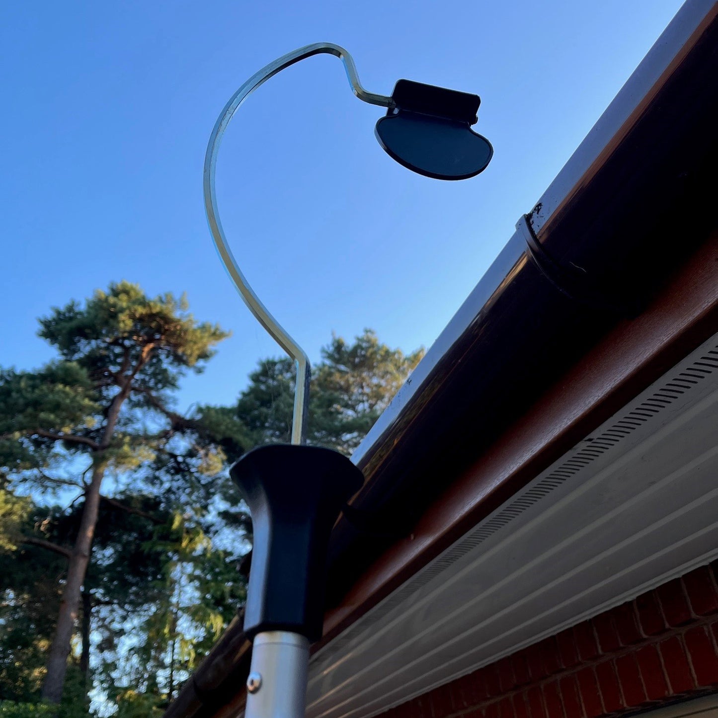 A lightweight gutter cleaner with a black paddle attached to a silver pole, shown in use against a domestic gutter backdrop.