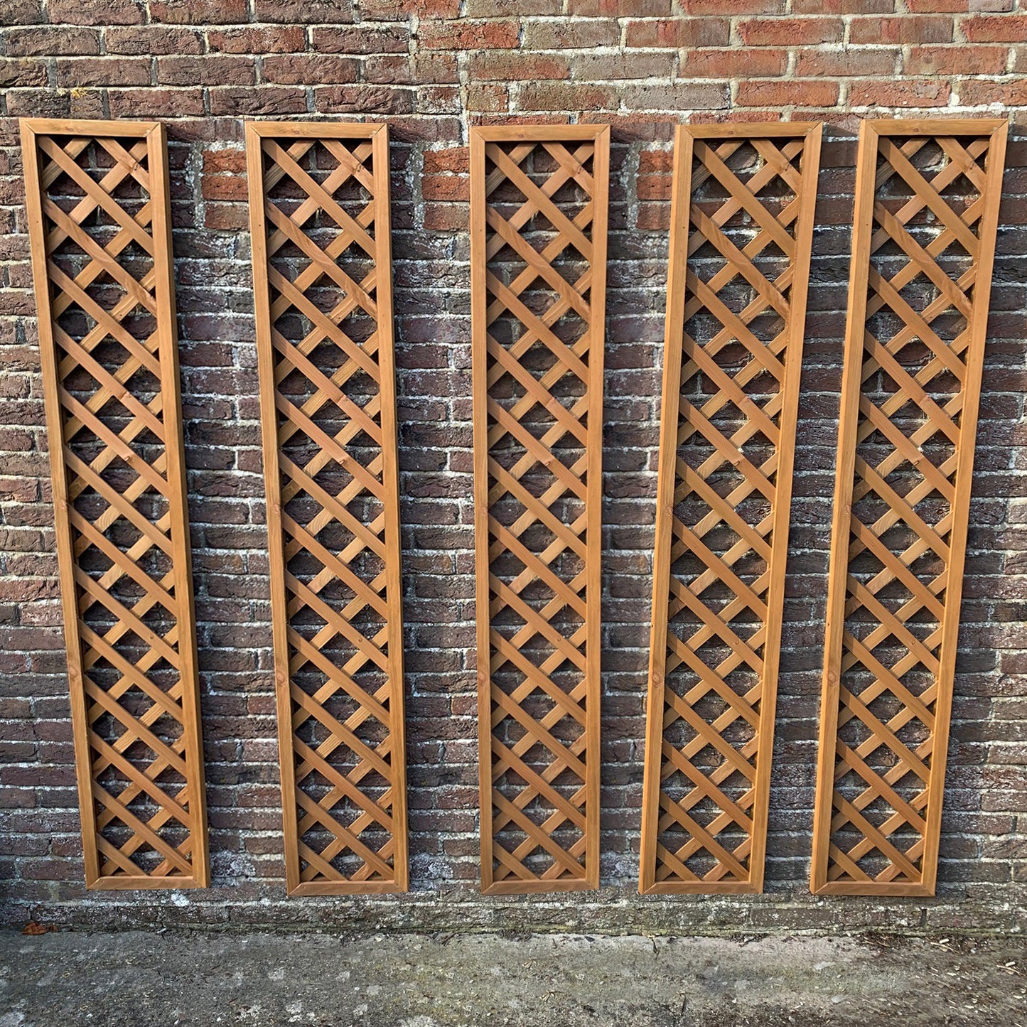 A set of five wooden framed trellis panels standing vertically against a brick wall.