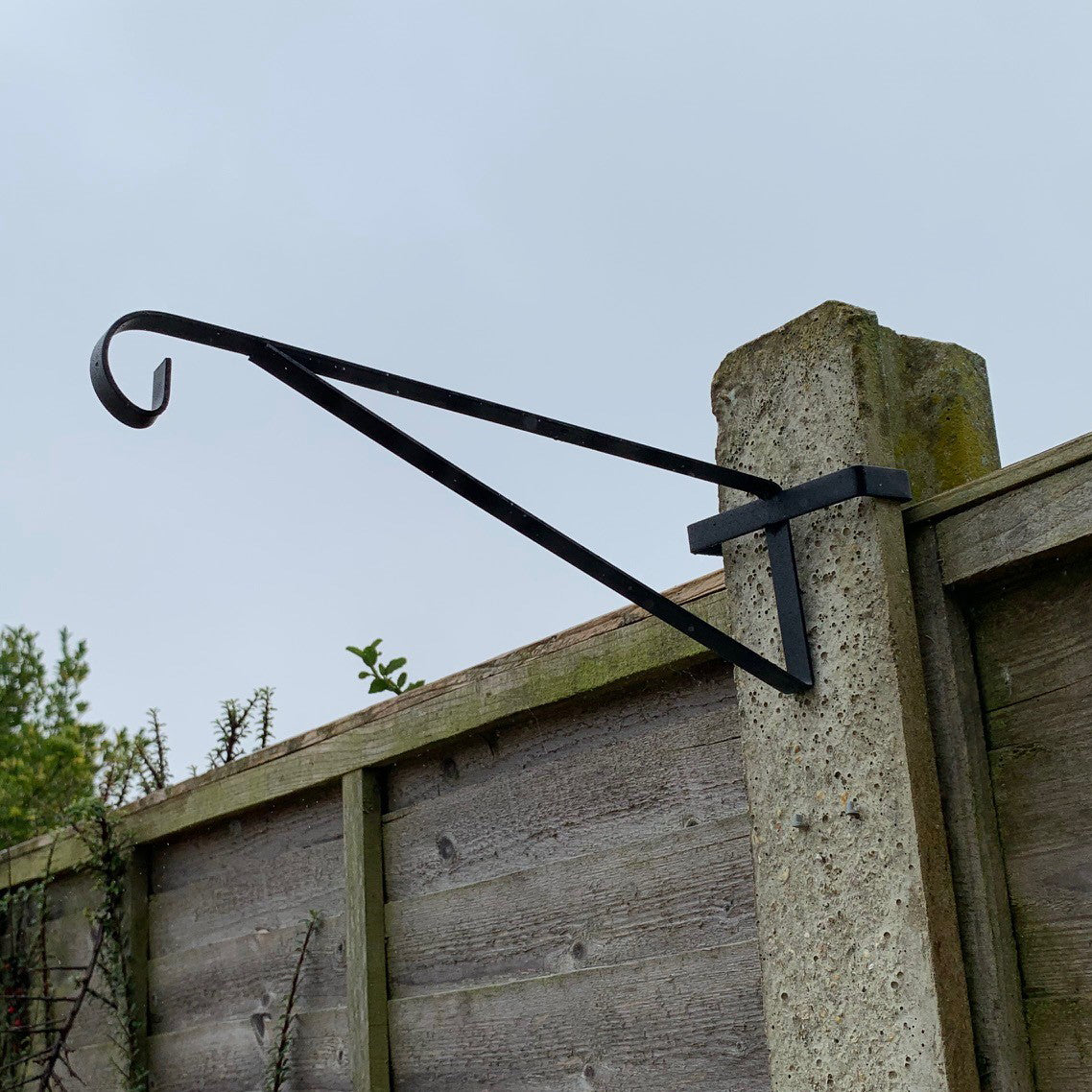 A black painted steel hanging basket bracket attached to a concrete fence post.