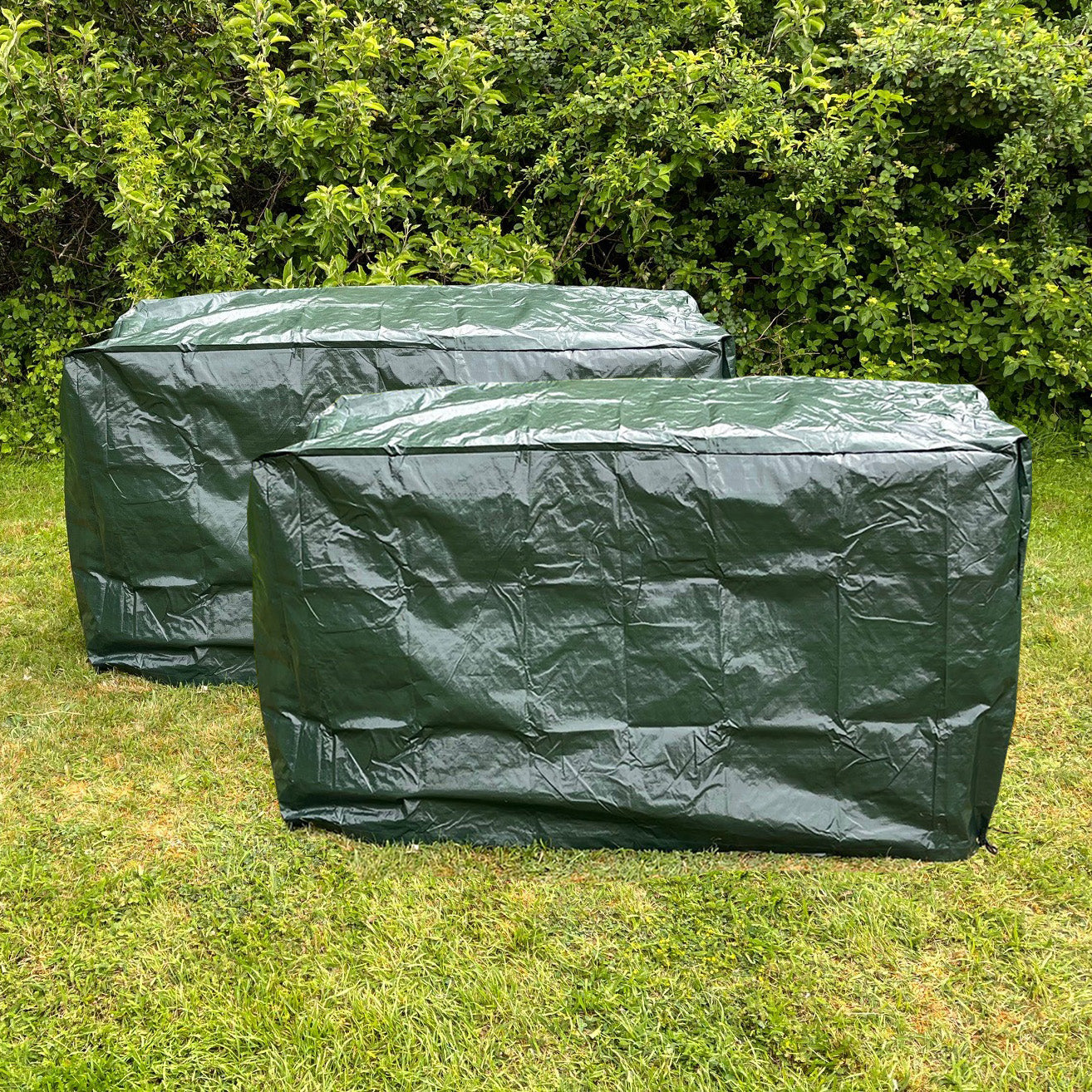 Two green waterproof garden bench covers on a grassy area with foliage in the background.