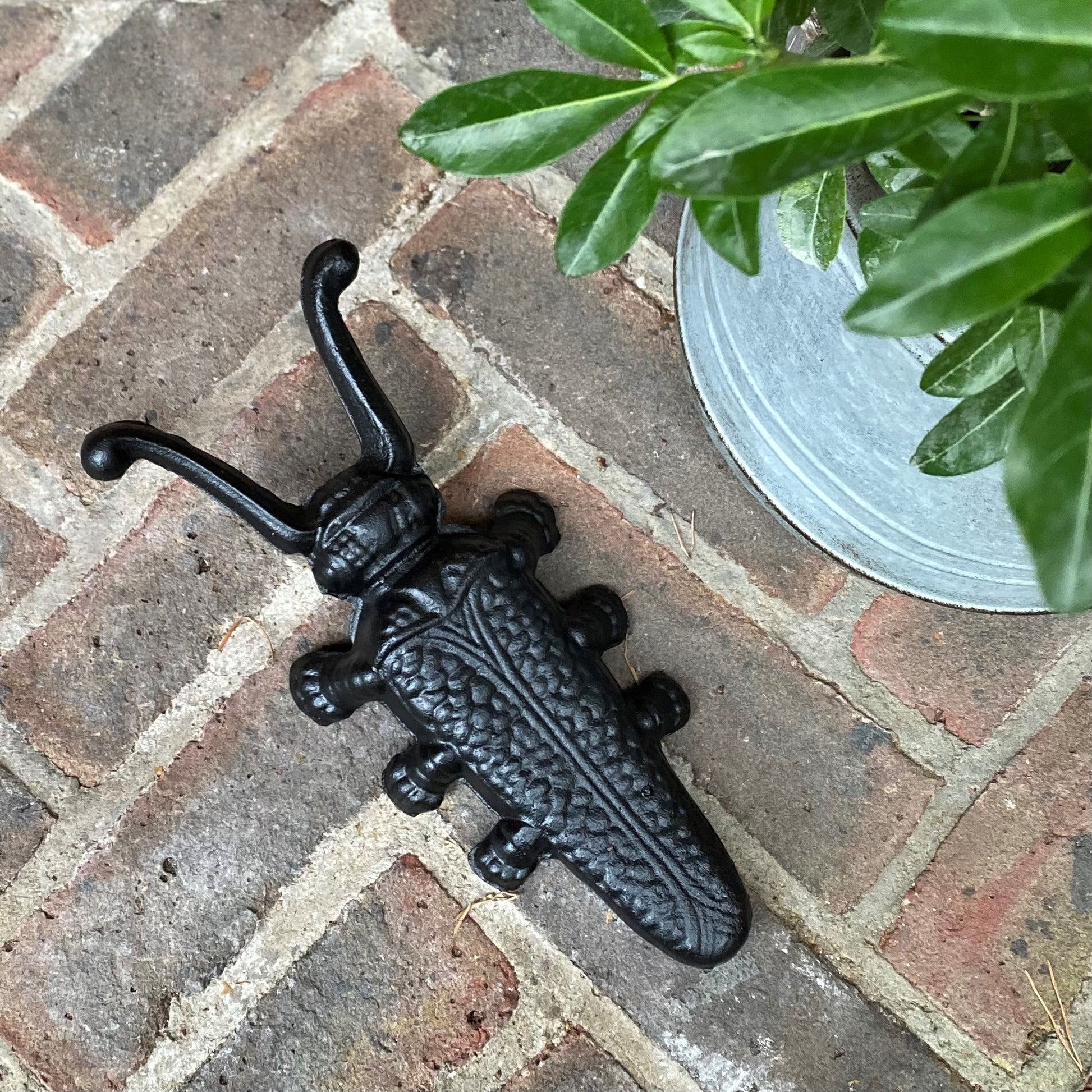A black cast iron boot jack designed to look like a beetle, placed on a brick surface with a plant in the background.