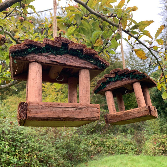 Two rustic style wooden hanging bird table feeders attached to a tree branch, set against a backdrop of green foliage.