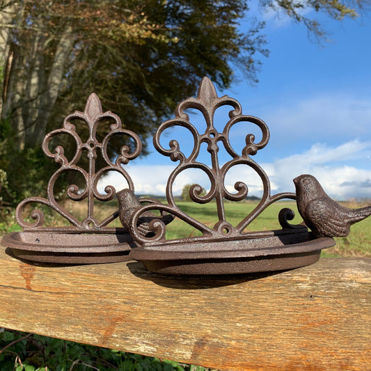 Two brown cast iron wall mounted bird feeders with decorative back panels and birds perched on the side, set against a wooden background.