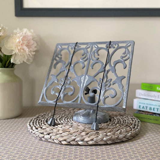 A traditional grey cast iron cookbook stand with an ornate floral pattern, displayed on a table.