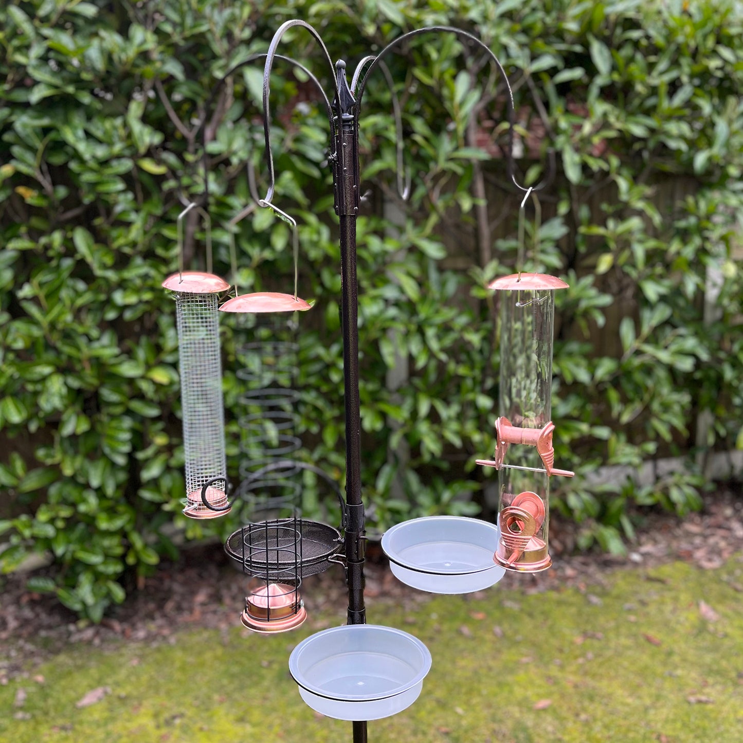 A metal bird feeding station with three copper style feeders hanging from a black pole, set against a green, leafy background.