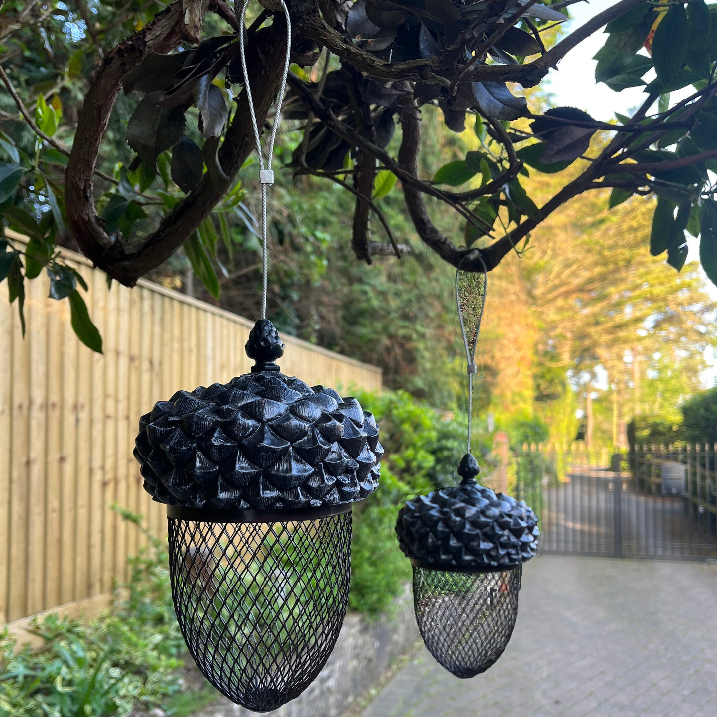 Two acorn-shaped bird feeders with a bronze effect, hanging from wires against a backdrop of green leaves and a wooden fence.
