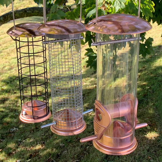Three copper bird feeders of different designs hanging outdoors on a grassy background.