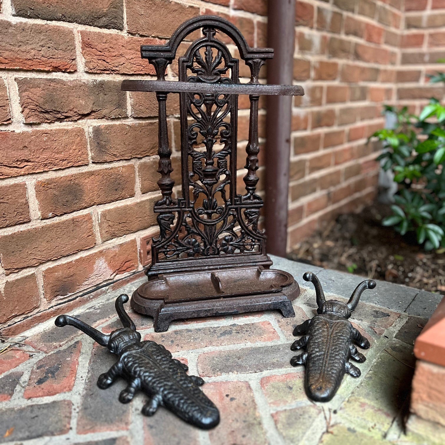 A brown cast iron umbrella stand with two black cast iron beetle boot jacks placed on the ground beside it, against a brick wall background.