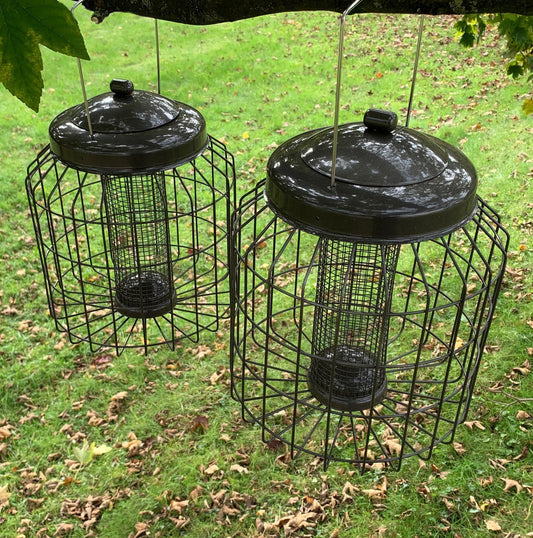 Two gun metal grey squirrel proof hanging bird nut feeders set on grass with leaves around.