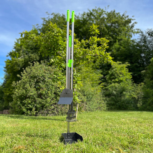 A set of green and silver dog pooper scooper tools placed on grass, including a rake, a spade, and a tray with long handles.