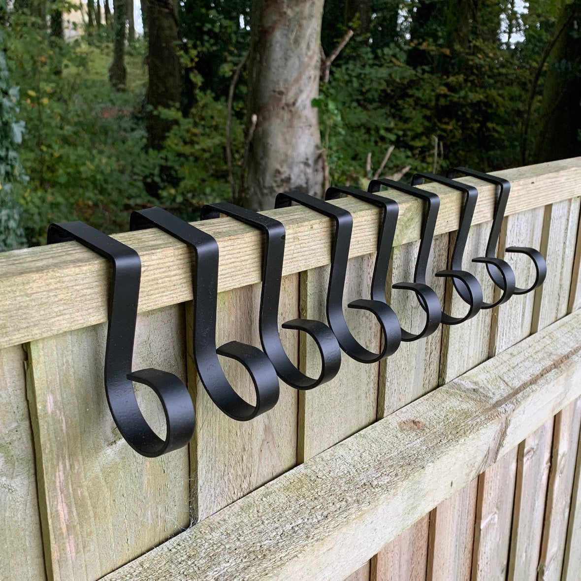 A set of black painted steel hooks installed on a wooden fence, designed to hang plants or decorations.