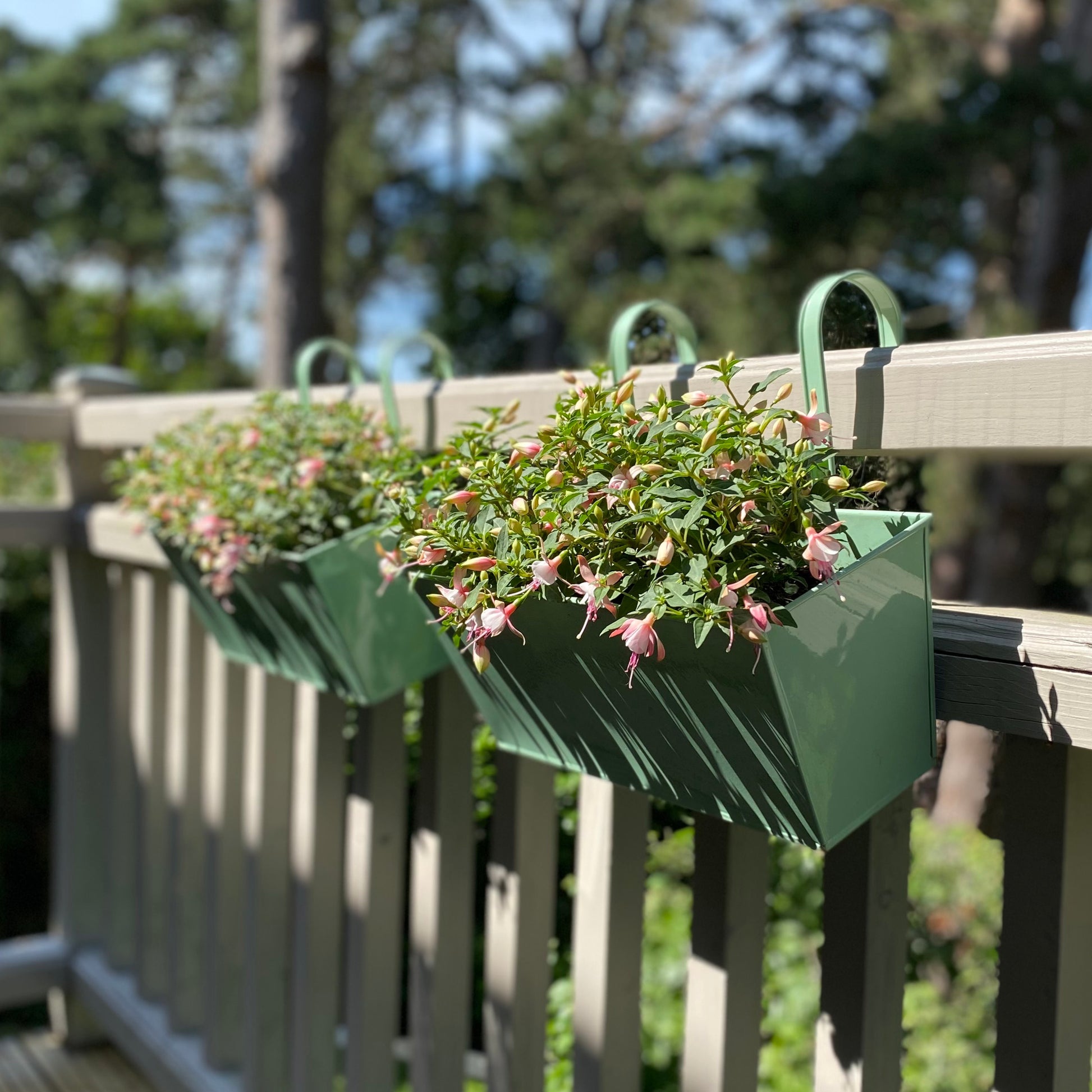 Two green metal balcony hanging planters with plants, attached to a wooden railing.