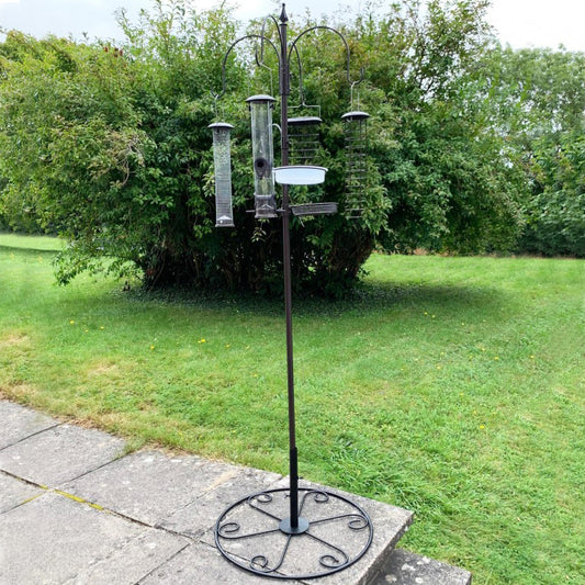 A metal bird feeding station with four large feeders mounted on a patio stand, positioned on a grassy area with trees in the background.