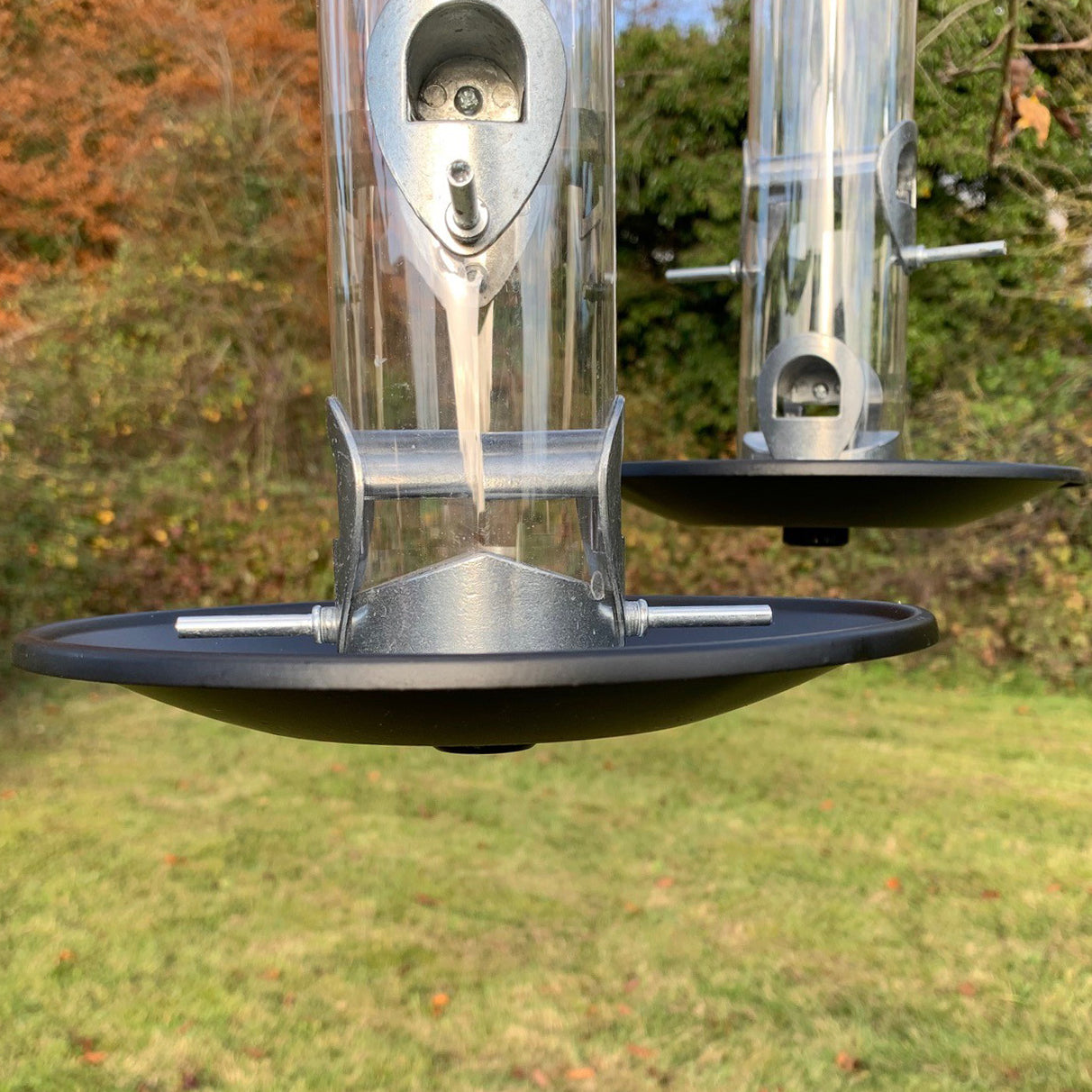 Two black seed catcher trays attached to bird feeders with a blurred natural background.