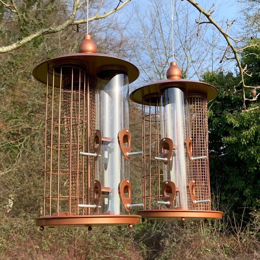 Two metal bird feeders designed for seeds, nuts, and fatball, with a copper finish and hanging loops, set against a natural background.