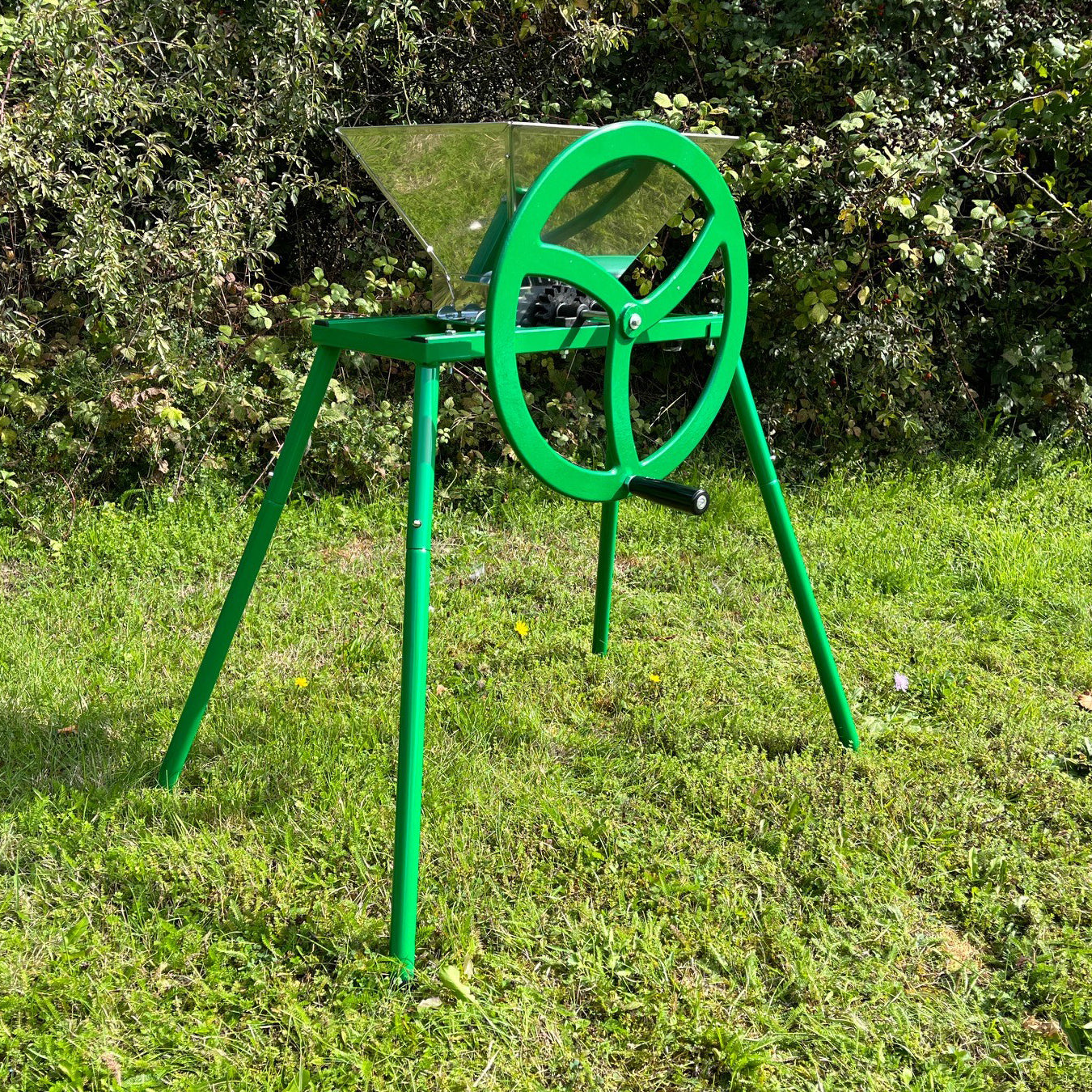 A green manual apple scratter pulper with a stand placed on grass against a shrubbery background.