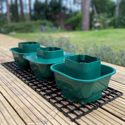 Three green plastic growbag watering pots placed on a black interlocking plastic mat, set outdoors on a wooden surface.