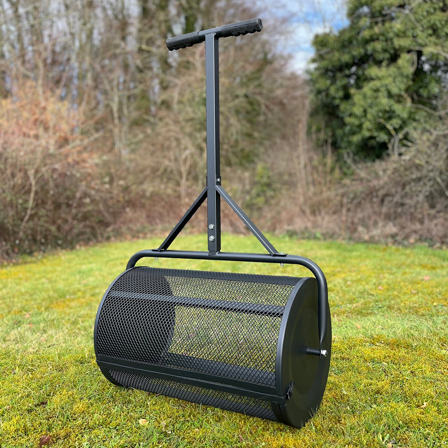 A black metal rolling garden lawn compost spreader with a mesh drum, standing on grass.