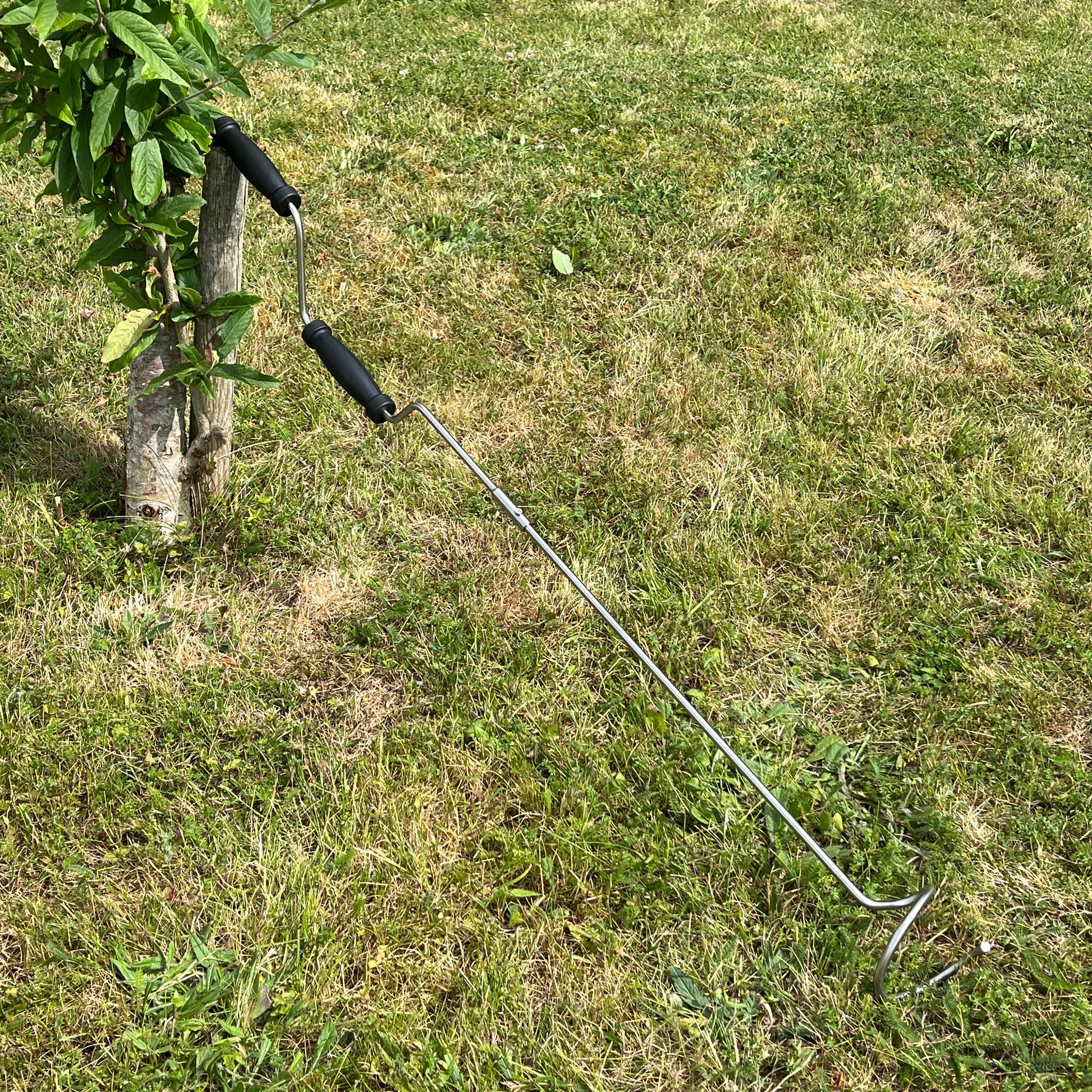 A metal compost aerator crank with plastic handles, placed on a grassy lawn.