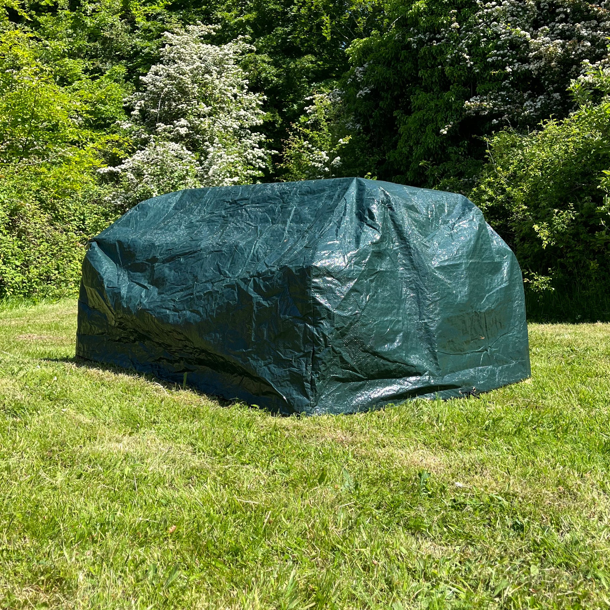 A green waterproof picnic table garden furniture cover placed on a grassy area, with trees and shrubs in the background.