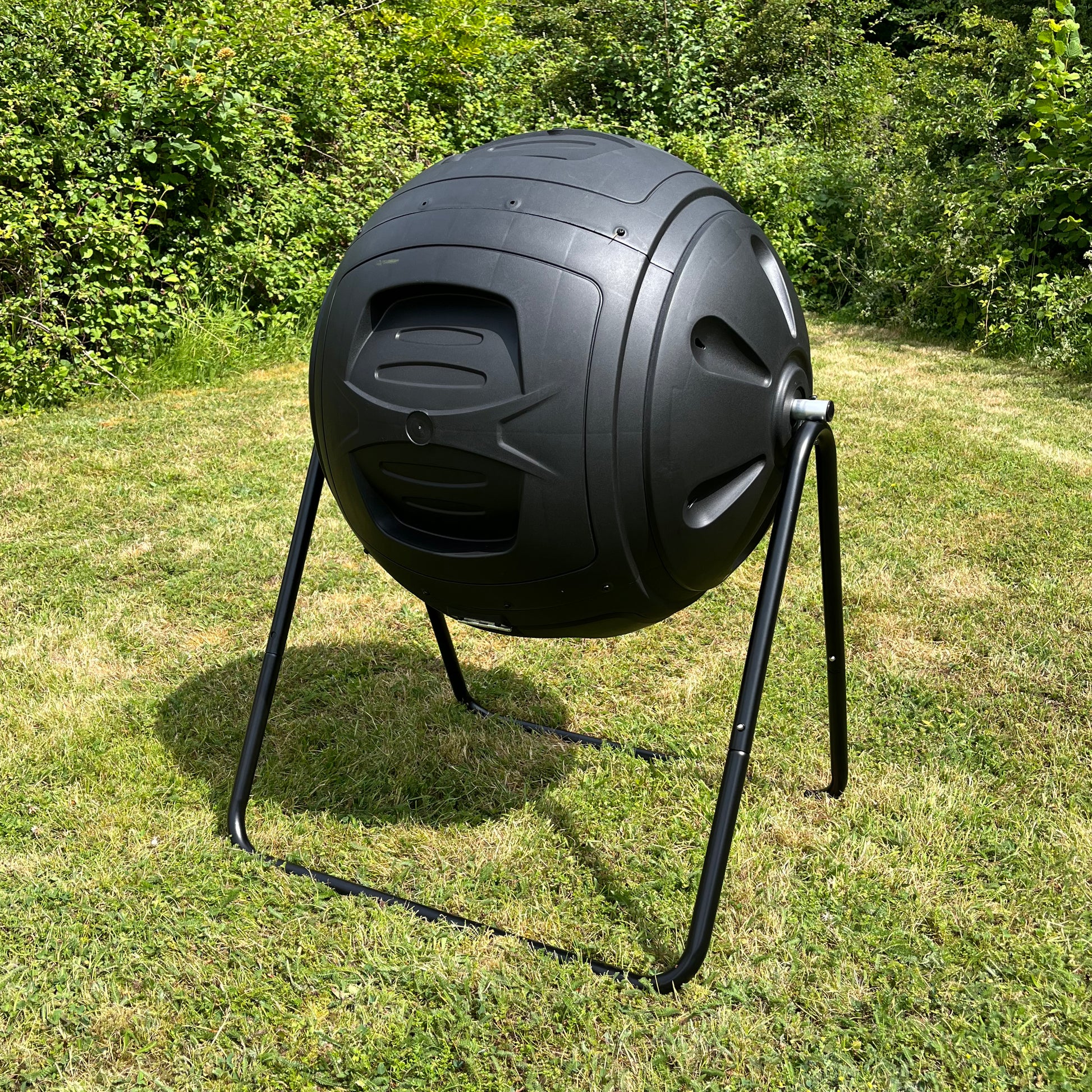 A black heavy duty garden compost tumbler placed on grass, designed with a horizontal rotating drum and a push-button lid locking mechanism.