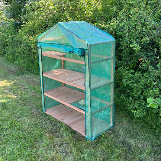 An extra wide 4 tier wooden mini greenhouse with a reinforced cover, placed outdoors on a grassy area beside some shrubbery.