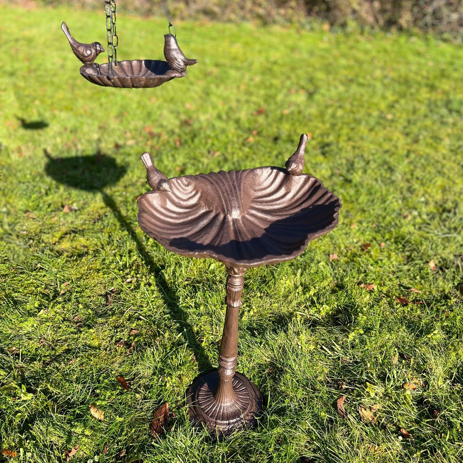A bronze-colored cast iron bird bath set with a decorative stand and hanging bowl, displayed on a grassy surface.