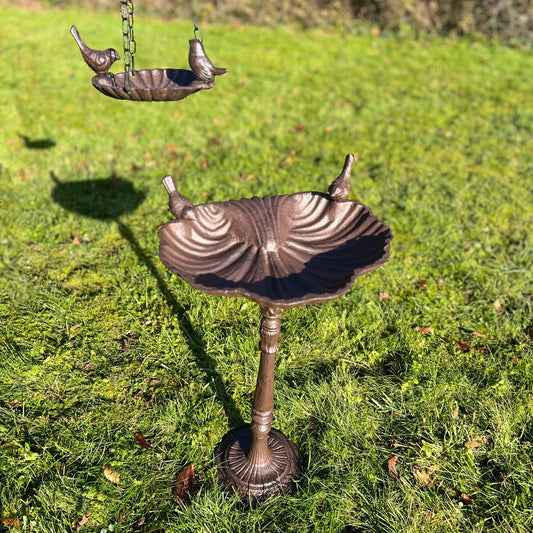 A bronze-colored cast iron bird bath set with a decorative stand and hanging bowl, displayed on a grassy surface.
