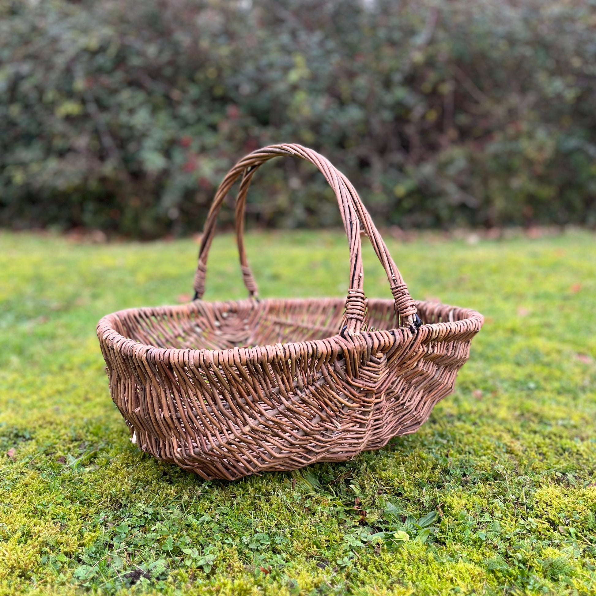 A large natural willow wicker garden trug basket with folding handles placed on a grassy surface.