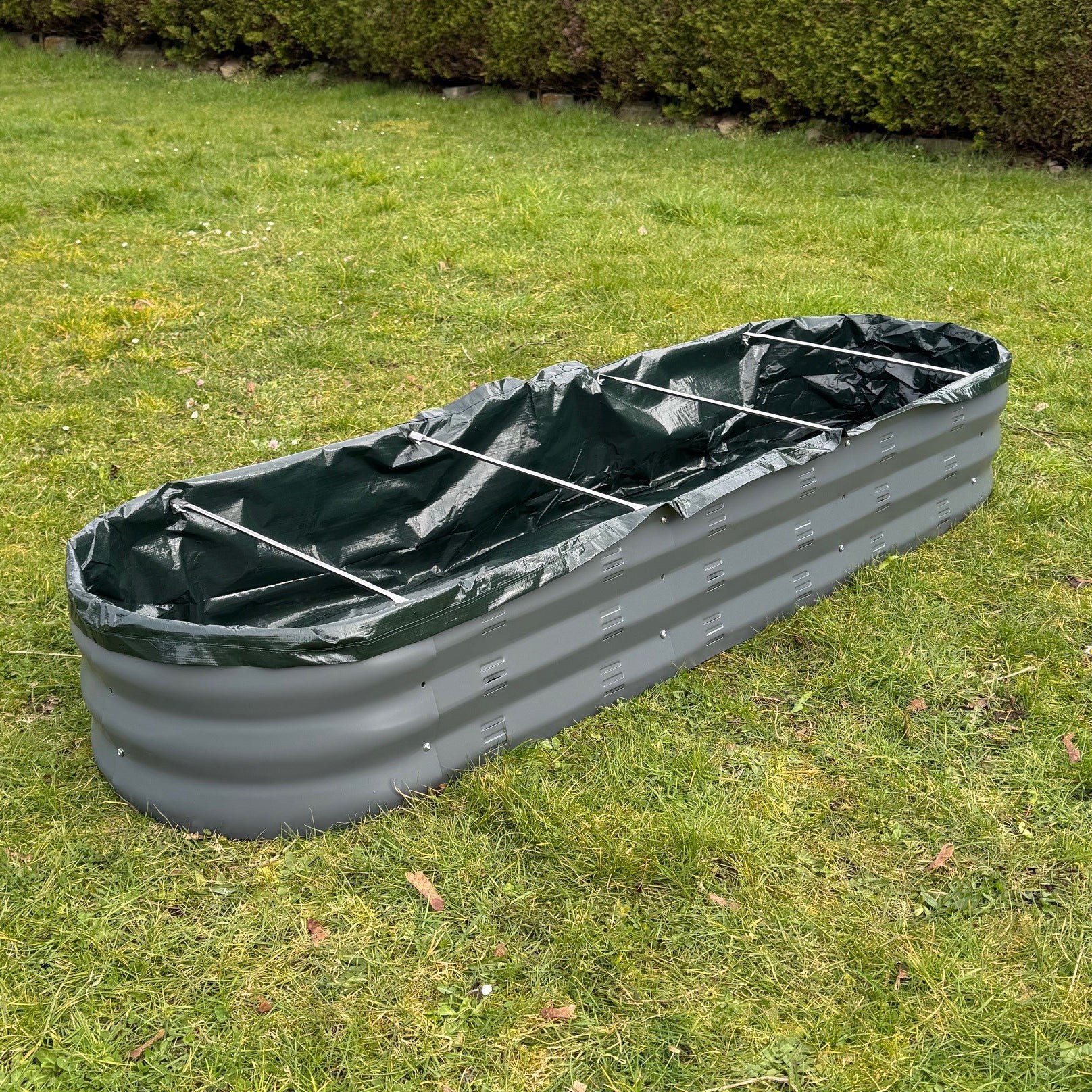A green liner for an oval metal raised vegetable bed laid on grass.
