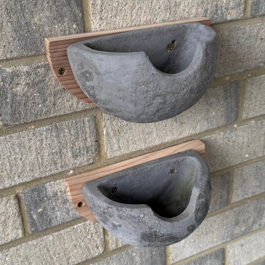 Two concrete bird nest boxes with wooden backing, mounted on a brick wall, with pre-drilled holes visible.