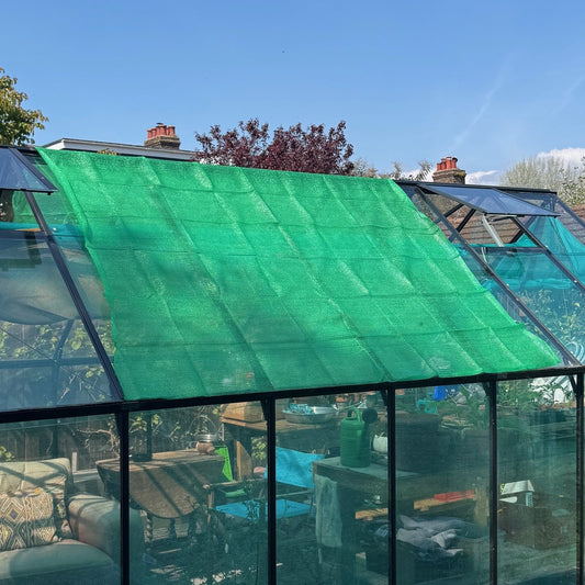 A green mesh sun shade and privacy screen net installed on a greenhouse, covering the glass panels.